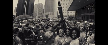 Movie still from “Battle of the Sexes” (2017), directed by Valerie Faris – A group of people holding up a microphone in a crowd; Extreme Wide shot, High angle