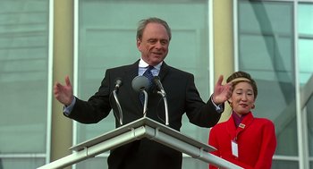 Movie still from “Bean” (1997), directed by Mel Smith – A man standing at a podium with microphones in front of a woman; Medium shot, Low angle