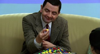 Movie still from “Bean” (1997), directed by Mel Smith – A man sitting on a couch with a tray of candy; Close Up shot, Over the shoulder angle