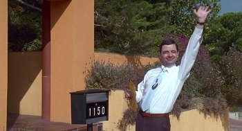 Movie still from “Bean” (1997), directed by Mel Smith – A man standing next to a mailbox holding a clipboard; Medium shot, Low angle