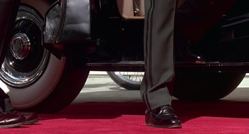 Movie still from “Bean” (1997), directed by Mel Smith – A man standing next to a car on a red carpet; Wide shot, Low angle