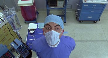 Movie still from “Bean” (1997), directed by Mel Smith – A man wearing a surgical mask in a room with other medical equipment; Close Up shot, Overhead angle