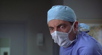 Movie still from “Bean” (1997), directed by Mel Smith – A man wearing a surgical mask and a blue surgical cap; Close Up shot, Low angle