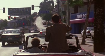 Movie still from “Bean” (1997), directed by Mel Smith – A man driving a car while another man holds his hand up to his ear; Wide shot, Over the shoulder angle