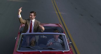 Movie still from “Bean” (1997), directed by Mel Smith – A man driving a red car with a dog in the back seat; Wide shot, High angle
