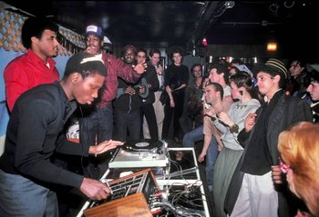 Movie still from “Beastie Boys Story” (2020), directed by Spike Jonze – A group of people standing around a table with a record player on top of it; Wide shot, Over the shoulder angle