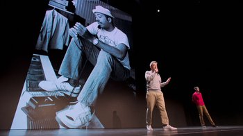 Movie still from “Beastie Boys Story” (2020), directed by Spike Jonze – A man standing in front of an image of a man on stage; Wide shot, Low angle
