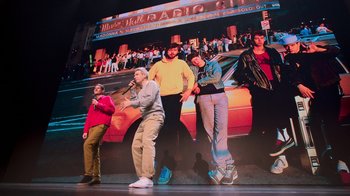 Movie still from “Beastie Boys Story” (2020), directed by Spike Jonze – A group of people standing in front of an image of a car; Wide shot, Low angle
