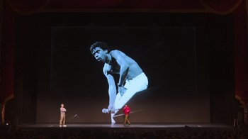 Movie still from “Beastie Boys Story” (2020), directed by Spike Jonze – A man standing in front of an image projected on a stage; Wide shot, Low angle