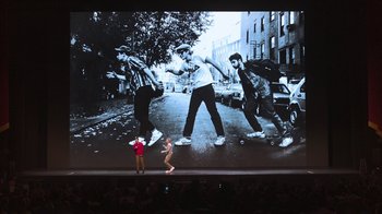 Movie still from “Beastie Boys Story” (2020), directed by Spike Jonze – A man standing in front of an image of a group of skateboarders; Wide shot, Low angle