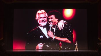 Movie still from “Beastie Boys Story” (2020), directed by Spike Jonze – A man standing in front of a large screen with an image of two men; Extreme Wide shot, Low angle