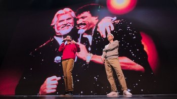 Movie still from “Beastie Boys Story” (2020), directed by Spike Jonze – Two men standing in front of an image of a man and a woman singing together; Wide shot, Low angle