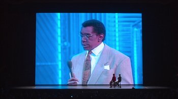 Movie still from “Beastie Boys Story” (2020), directed by Spike Jonze – A man in a suit and tie holding a microphone on a stage; Wide shot, Low angle