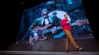 Movie still from “Beastie Boys Story” (2020), directed by Spike Jonze – A man standing in front of a large picture of a man playing a guitar; Wide shot, Low angle