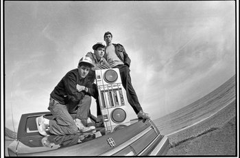 Movie still from “Beastie Boys Story” (2020), directed by Spike Jonze – Three young men sitting on top of a car with a boombox; Medium shot, Low angle