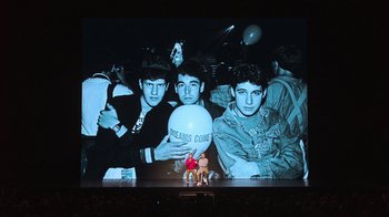 Movie still from “Beastie Boys Story” (2020), directed by Spike Jonze – A group of people sitting on a stage in front of a large screen; Wide shot, Low angle