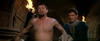 Movie still from “Becket” (1964), directed by Peter Glenville – A man with a beard is standing in front of a fire; Medium shot, Over the shoulder angle