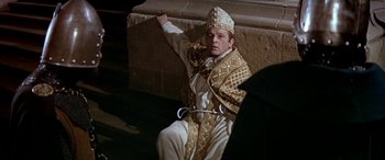 Movie still from “Becket” (1964), directed by Peter Glenville – A man wearing a white and gold outfit sitting next to a stone wall; Medium shot, High angle