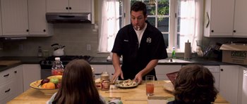 Movie still from “Bedtime Stories” (2008), directed by Adam Shankman – A man standing at a table with a plate of food in front of him; Medium shot, Over the shoulder angle