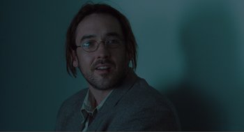 Movie still from “Being John Malkovich” (1999), directed by Spike Jonze – A man with long hair and glasses is wearing a suit; Close Up shot, Low angle