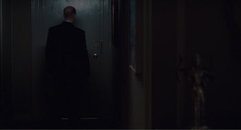 Movie still from “Being John Malkovich” (1999), directed by Spike Jonze – A man standing in a dark room next to a door; Wide shot, Over the shoulder angle