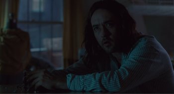 Movie still from “Being John Malkovich” (1999), directed by Spike Jonze – A man with long hair sitting in front of a window at night; Medium shot, Low angle
