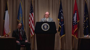 Movie still from “Being There” (1979), directed by Hal Ashby – A man standing at a podium in front of two flags; Wide shot, Over the shoulder angle