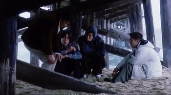 Movie still from “Better Luck Tomorrow” (2002), directed by Justin Lin – A group of people sitting on the sand under a bridge; Wide shot, High angle