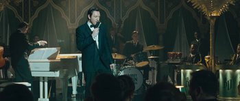 Movie still from “Beyond the Sea” (2004), directed by Kevin Spacey – A man in a suit is singing into a microphone in front of a group of people; Medium shot, Over the shoulder angle