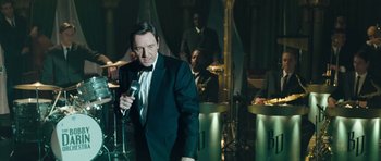 Movie still from “Beyond the Sea” (2004), directed by Kevin Spacey – A man in a tuxedo holding a microphone in front of a group of people; Medium shot, Over the shoulder angle