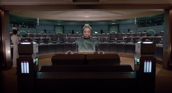 Movie still from “Bicentennial Man” (1999), directed by Chris Columbus – A woman sitting in front of an audience in front of a row of chairs; Medium shot, Low angle
