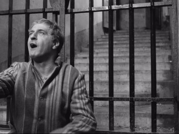 Movie still from “Big Deal on Madonna Street” (1958), directed by Mario Monicelli – A black and white photo of a man standing in front of a metal gate; Medium shot, Low angle