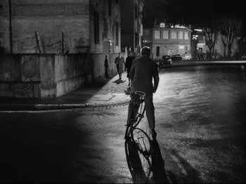 Movie still from “Big Deal on Madonna Street” (1958), directed by Mario Monicelli – A man riding a bike down a street at night; Wide shot, Over the shoulder angle