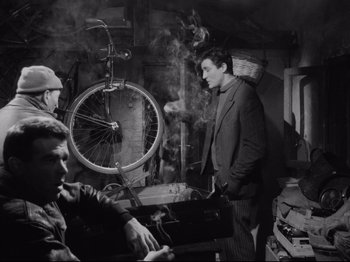 Movie still from “Big Deal on Madonna Street” (1958), directed by Mario Monicelli – A black and white photo of a man standing in front of a bicycle wheel; Medium shot, High angle