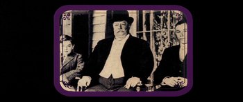 Movie still from “Big Jake” (1971), directed by George Sherman – An old photo of a man in a top hat sitting next to two other men; Medium shot, Low angle