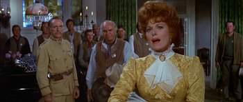 Movie still from “Big Jake” (1971), directed by George Sherman – A group of people standing in a room with a woman in a yellow dress; Medium shot, Over the shoulder angle