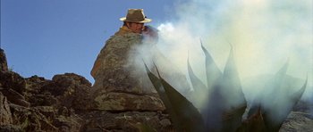 Movie still from “Big Jake” (1971), directed by George Sherman – A man in a hat is smoking a pipe on top of a stone wall; Medium shot, Low angle