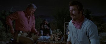 Movie still from “Big Jake” (1971), directed by George Sherman – A group of people sitting around a table at night; Medium shot, Over the shoulder angle