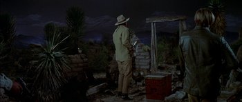 Movie still from “Big Jake” (1971), directed by George Sherman – A man in a hat standing next to a wooden structure; Wide shot, Low angle
