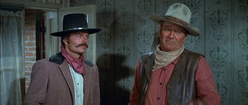 Movie still from “Big Jake” (1971), directed by George Sherman – A couple of men standing next to each other in a room; Medium shot, Over the shoulder angle