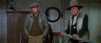 Movie still from “Big Jake” (1971), directed by George Sherman – A man and a woman standing next to each other in a room; Medium shot, Over the shoulder angle