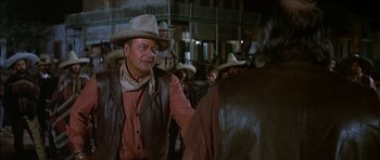 Movie still from “Big Jake” (1971), directed by George Sherman – An older man wearing a cowboy hat in front of a group of people; Medium shot, Over the shoulder angle