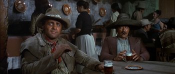 Movie still from “Big Jake” (1971), directed by George Sherman – Two men sitting at a table with a beer in front of them; Medium shot, Low angle
