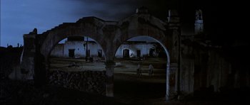 Movie still from “Big Jake” (1971), directed by George Sherman – Two people riding bikes through an archway in a dark place; Extreme Wide shot, Low angle