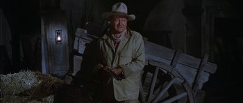 Movie still from “Big Jake” (1971), directed by George Sherman – An older man in a cowboy hat holding a gun in front of an old wagon; Medium shot, Over the shoulder angle