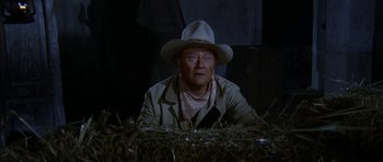 Movie still from “Big Jake” (1971), directed by George Sherman – An older man wearing a cowboy hat sitting next to hay bales; Medium shot, Over the shoulder angle