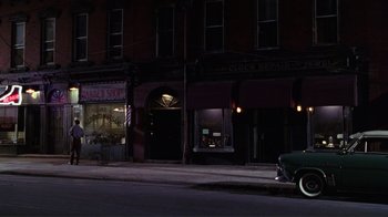 Movie still from “Big Night” (1996), directed by Stanley Tucci – A car parked on the side of the road in front of a barber shop at night; Wide shot, High angle