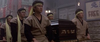 Movie still from “Big Trouble in Little China” (1986), directed by John Carpenter – Two men sitting in front of a casket in a room; Medium shot, Low angle