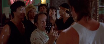Movie still from “Big Trouble in Little China” (1986), directed by John Carpenter – A group of people standing around a man holding a gun; Medium shot, Over the shoulder angle