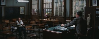 Movie still from “Bigger Than Life” (1956), directed by Nicholas Ray – A person sitting at a desk in a room with chairs; Wide shot, High angle